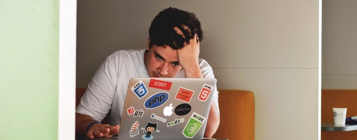 hombre sentado tocándose la cabeza con la mano mientras mira su computadora gris con stickers de colores; hombre con estrés por trabajo
