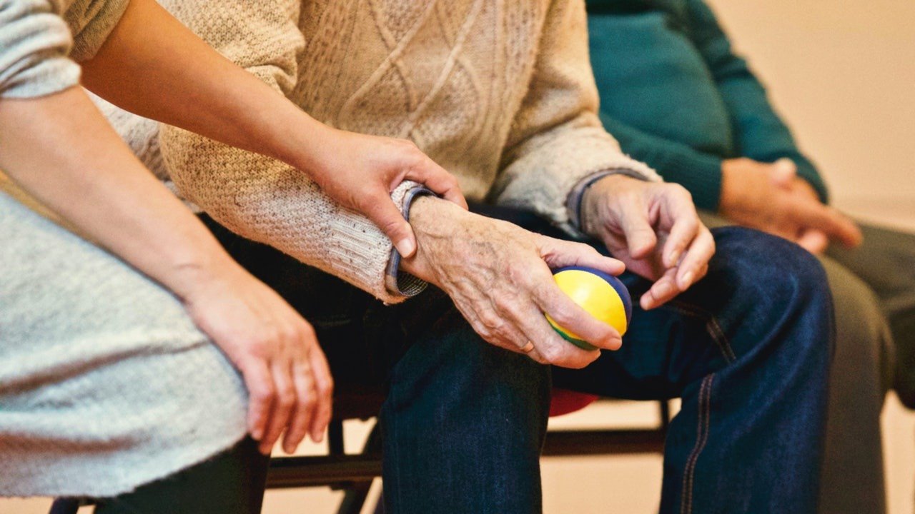 Mano de hombre joven  encima de una mano de una persona adulta que tiene suéter color crema, en su mano derecha tiene una pelota color amarilla; bienestar psicológico