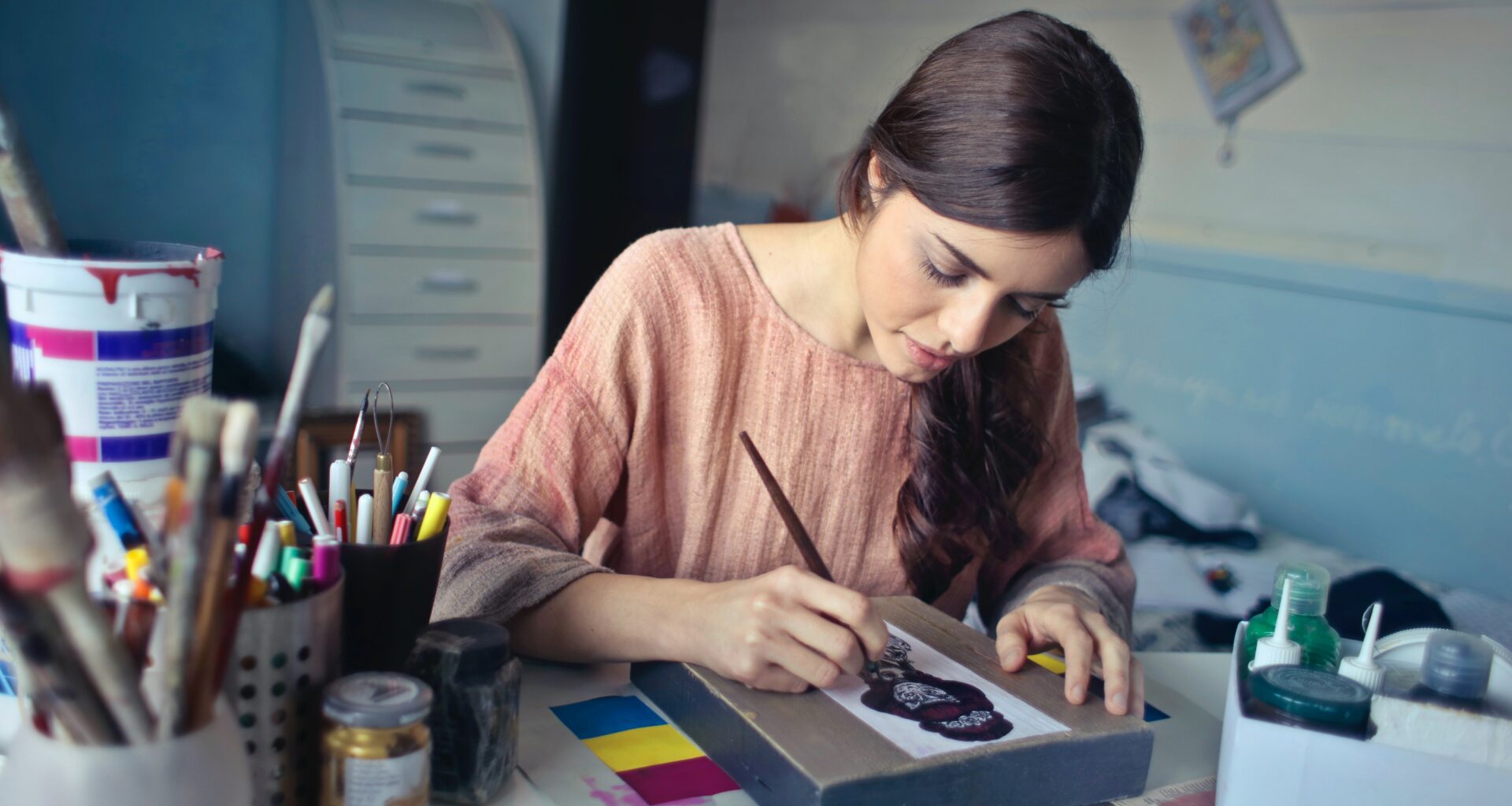 mujer de cabello largo y blusa rosa pinta con un pincel sobre un lienzo, a su lado hay colore sy pinturas dentro de un bote; diseño gráfico sustentable