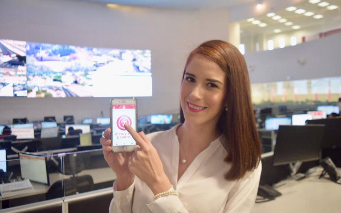 Cassandra López Manzano mostrando App Mujeres Seguras en su teléfono blanco. Detrás hay monitores de computadoras