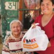 mujer de blusade manga larga roja sostiene bolsa de plástico con alimentos en el interior y a un costado una señora anciana sonríe