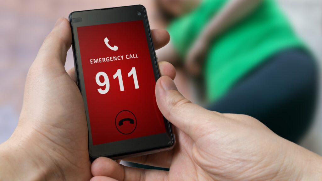 Manos con un celular color negro y la pantalla color rojo con numero de emergencia 911 con color blanco. La parte de arriba del numero con letras color blanco "EMERGENCY CALL", para localizar a migrantes perdidos en Sonora.