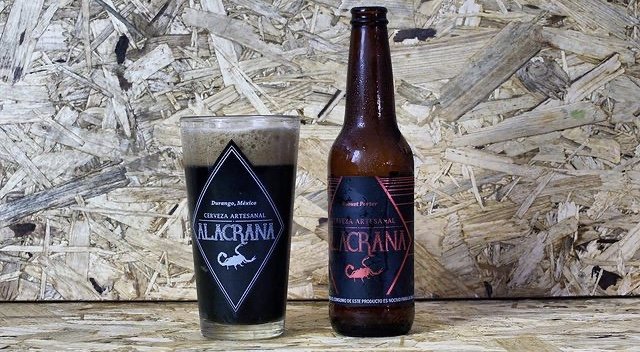 Vaso de cerveza con logotipo con letras que dicen Alacrana, a la derecha una botella de vidrio color ambar con etiqueta color negro, de fondo color madera clara.