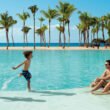 niña y niño corren a la orilla de la piscina donde etsán sentados dos adultos, detrás hay palmeras y el mar