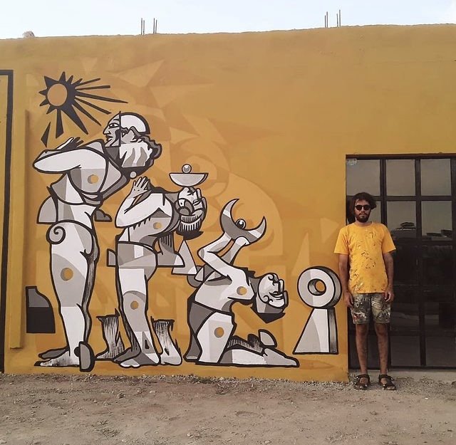 Joven con camisa color amarilla, short color negro, salpicado de pintura de diversos colores, detrás de el mural con tres figuras humanas de color blanco-gris y el fondo color amarillo