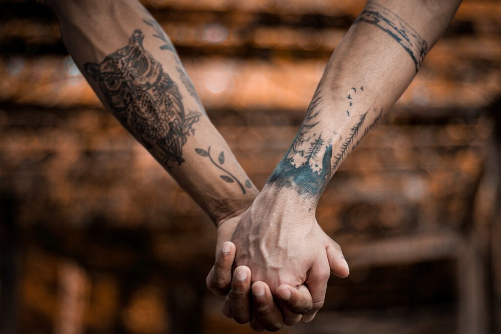 Manos con tatuajes enlazadas. La mano del lado izquierdo tiene un tatuaje con tinta negra de un búho y la mano derecha tiene tatuado con tinta negra un bosque con un lobo.