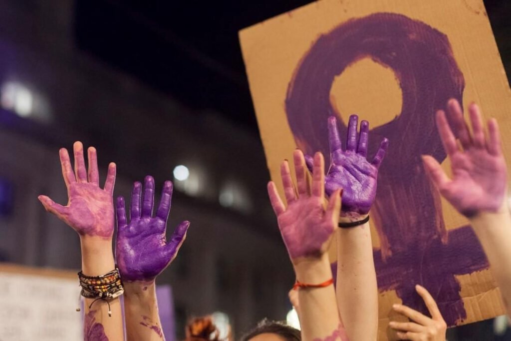Mujeres alzando manos pintadas con color morado, detras de estas manos un cartel de carton con el simbolo de sexo femenino color morado.