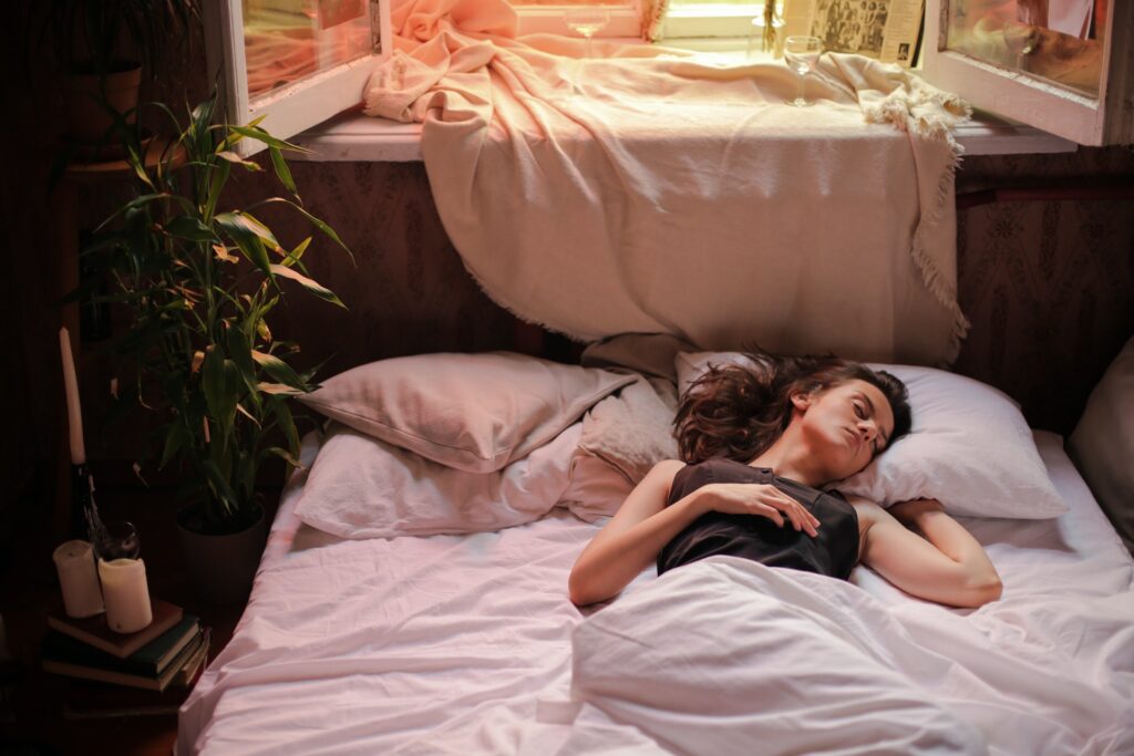 Mujer con blusa color negro, dormida en cama con sabanas color blancas. Del lado izquierdo se encuentra una planta grande junto a veladoras color blancas.