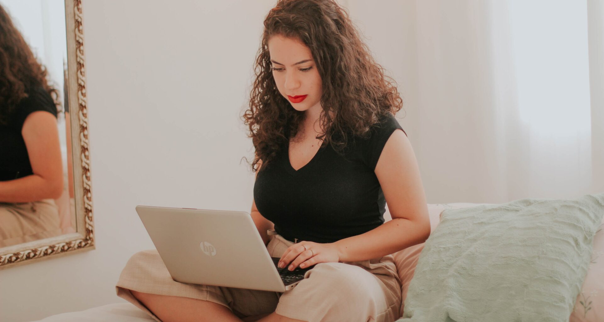 mujer con cabello rizado y labios pintados de rojo, blusa negra y pantalones claros, está sentada sobre la cama escribiendo en la computadora