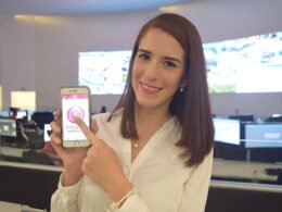 mujer de cabello lacio sonríe y sostiene un teléfono blanco mientras presiona la pantalla con su dedo índice; atrás hay monitores de computadoras