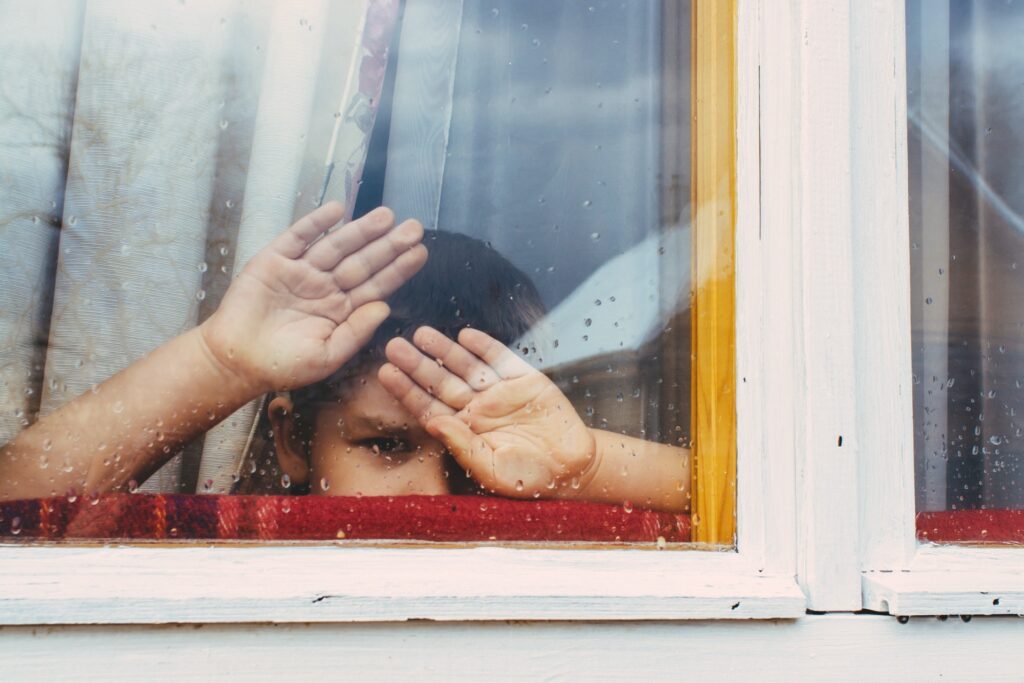 Niño con manos en ventana color blanca, el cristal con gotas de agua.