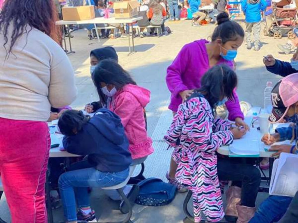 Niños y niñas migrantes sentados en mesas escribiendo. Del lado derecho se encuentra una niña parada con sueter morado y cubrebocas azul.