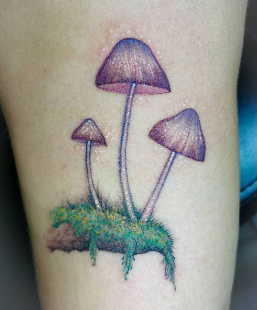 Tatuaje Hermosillo- Tatuaje de tres hongos color morado-rosa con puntos color blanco. Debajo de los hongos un pedazo de tierra con zacate y flores pequeñas color amarillo.