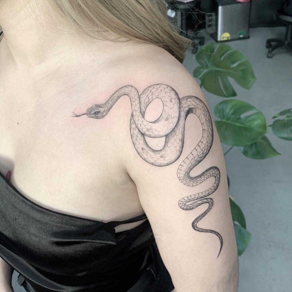 Hombro y brazo del lado izquierdo con un tatuaje con tinta negra de una serpiente.
