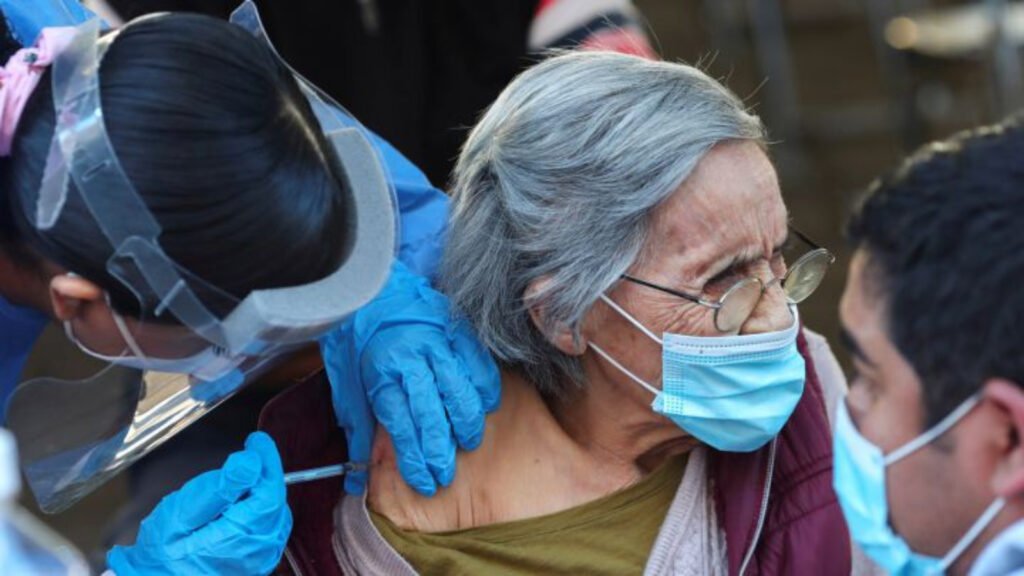 Señora adulta-mayor recibe vacunación en el brazo derecho. La señora tiene un cubrebocas color azul y anteojos. La persona que la esta vacunando tiene guantes color azul.