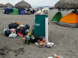 bote de basura verde con bolsas de plástico en el interior y fuera sobre la arena de la playa; detrás hay tiendas de acampar de colores bajo palapas