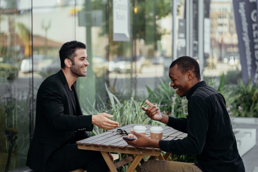 Informe mundial de la felicidad; dos hombres sentados en una mesa de madera riéndose. Los dos tienen camisa color negro y en la mesa se encuentran dos recipientes de café color blanco.