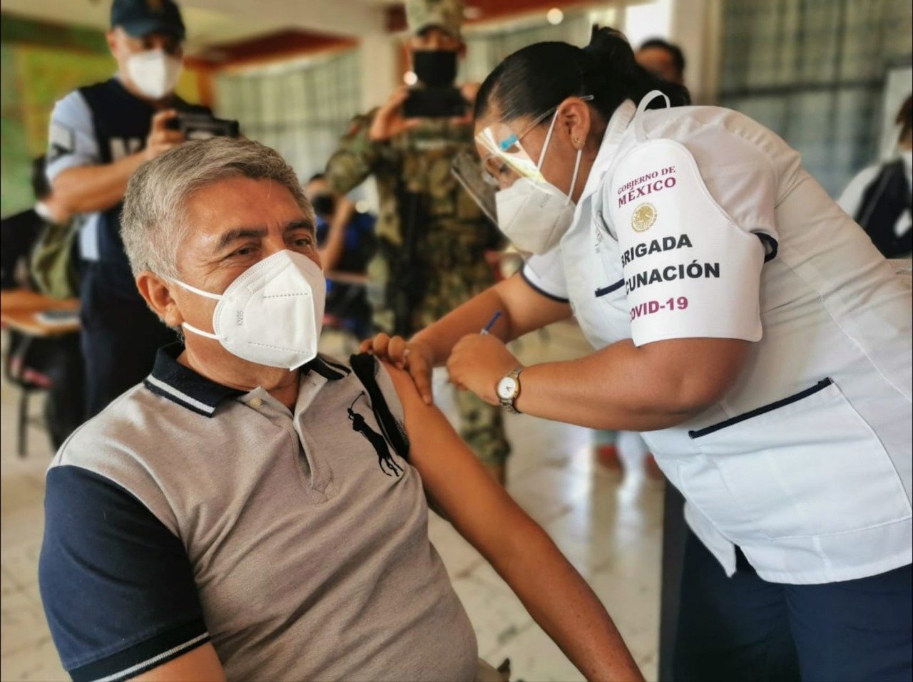 Señor adulto mayor con cubrebocas color blanco, camisa color gris claro con negro, recibe vacunacion de una señora que tiene filipina color blanco, careta y cubreboca color blanco.