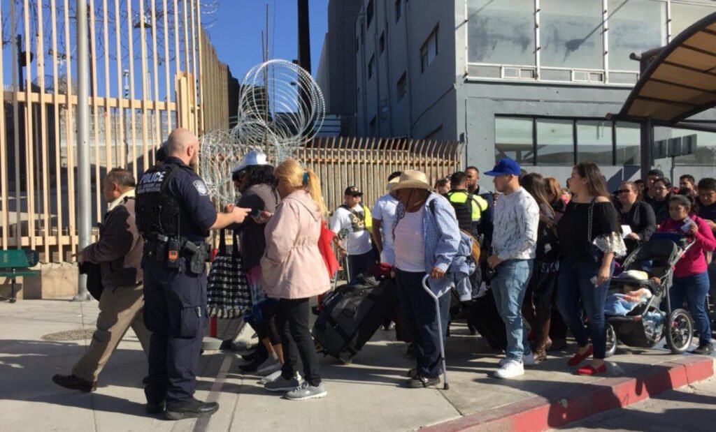 La frontera de Tijuana; se encuentran mujeres, hombres y niños y niñas haciendo fila para entrar a los EUA. En la parte del fondo se encuentra una reja de color crema y un edificio color gris.
Del lado izquierdo se encuentra un hombre con uniforme de policia color negro.