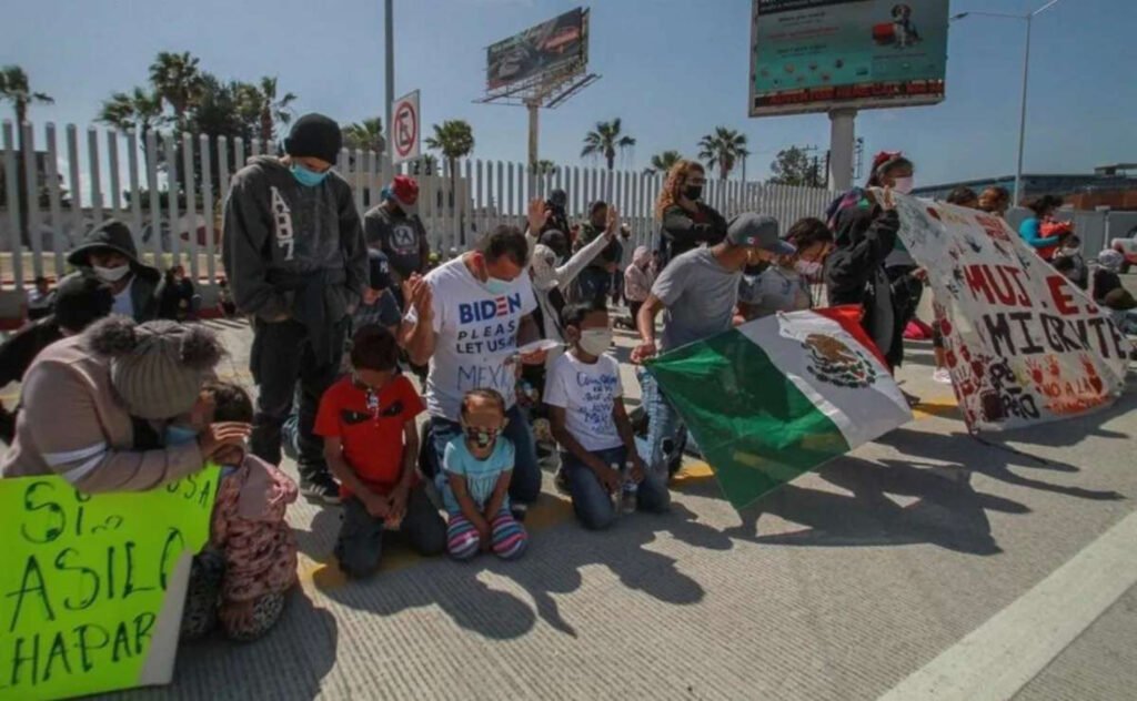 Migrantes se encuentran de rodillas en la linea. En medio un chico tiene una bandera mexicana color, verde, blanco y rojo y en medio una águila. 
Del lado izquierdo una mujer tiene una cartulina color fosforescente.
