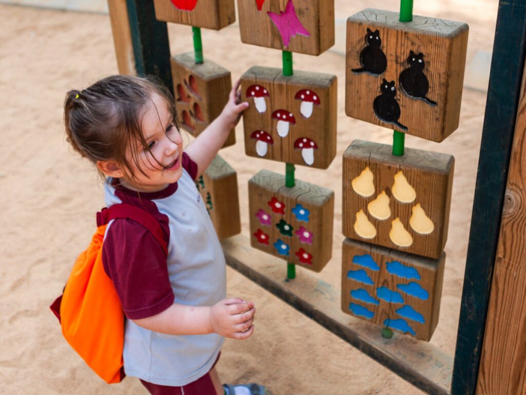 Sofía XT; niña con camisa color blanco con guinda, mochila color naranja, jugando con bloques de madera con diferentes figuras.