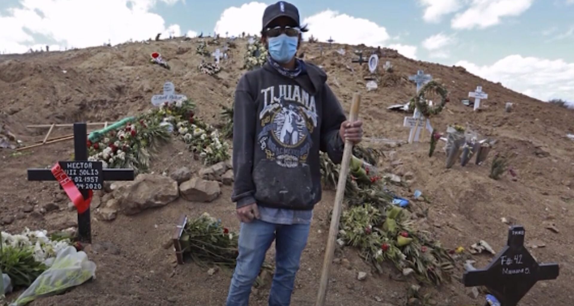 hombre con sudadera que dice Tijuana, gorra y cubrebocas sostiene un palo en medio del cementerio