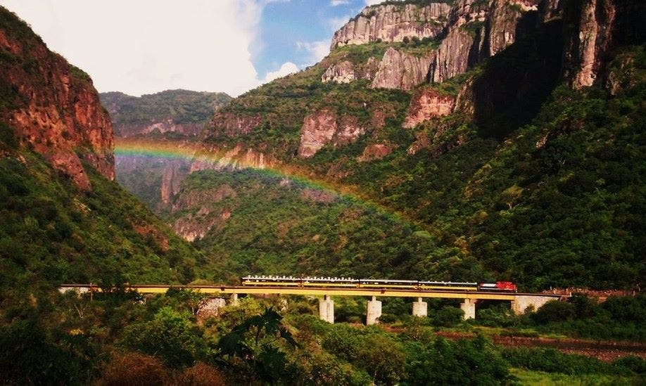Paisaje montañoso con el turistico tren del Chepe atravesando bajo el arcoiris en las Barrancas del Cobre en Chihuahua