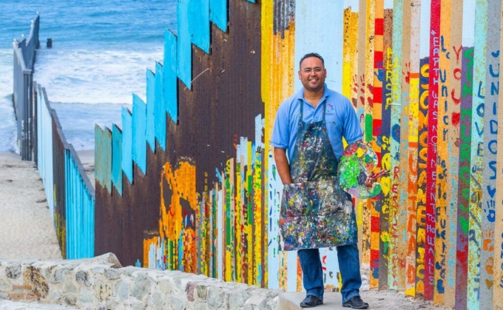 Foto del Mural de la Hermandad junto a su creador, el artista Enrique Chiu; este punto de Tijuana representa el límite que tiene Estados Unidos con México, y saldrá en la película "Welcome To al norte". FOTO: Portal 24 Horas.