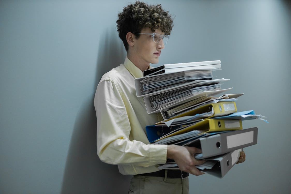 In the image you can see a young man wearing glasses. Her hair is long and curly. She wears a light yellow long shirt and khaki pants In her hands we can see some folders