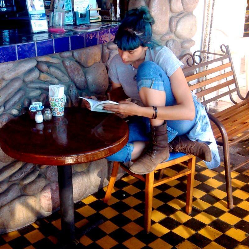 La psicóloga y artista Rofelda González sentada en una silla mientras lee un libro. Viste botas café; su cabello está pintado de azul.