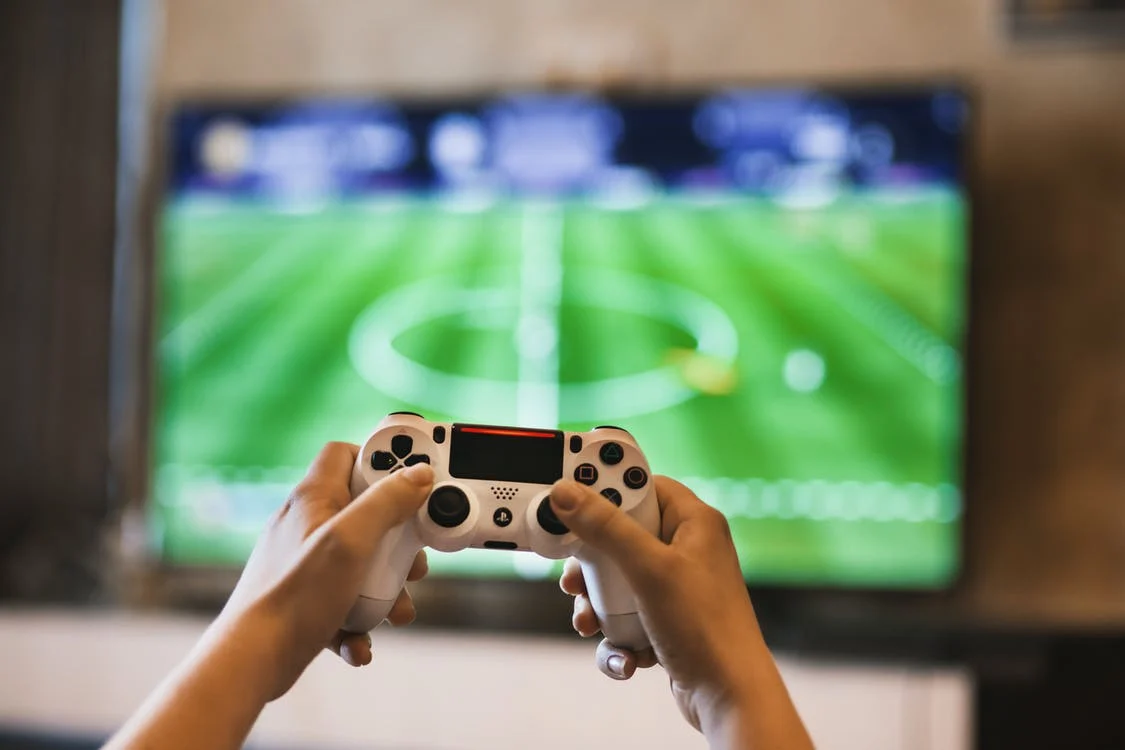 Un gamer tiene un control blanco de consola de video juego. De fondo hay una pantalla de televisión reproduce un campo de futbol.