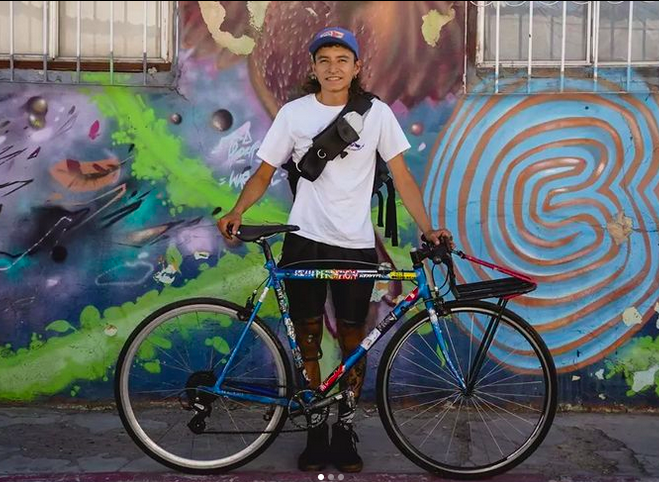Es uno de los ciclistas que desea recorrer el país en bicicleta. Es un hombre con gorra que posa directamente hacia la cámara, sostiene su bicicleta.