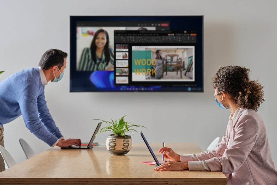 Un hombre y una mujer están en una conferencia virtual. Se recargan en una mesa, frente a sí una pantalla que emula estar en una situación de trabajo híbrido.