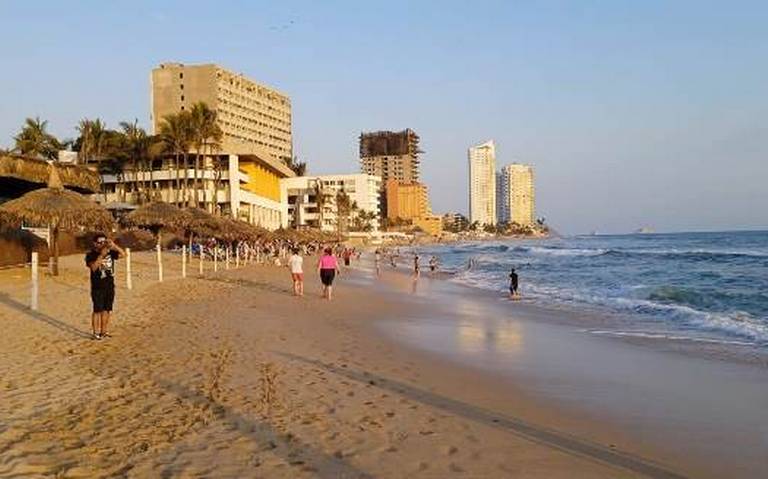 Es la imagen de la playa: arena del lado izquierdo y océano del derecho. Al fondo unos hoteles y algunas personas caminando.