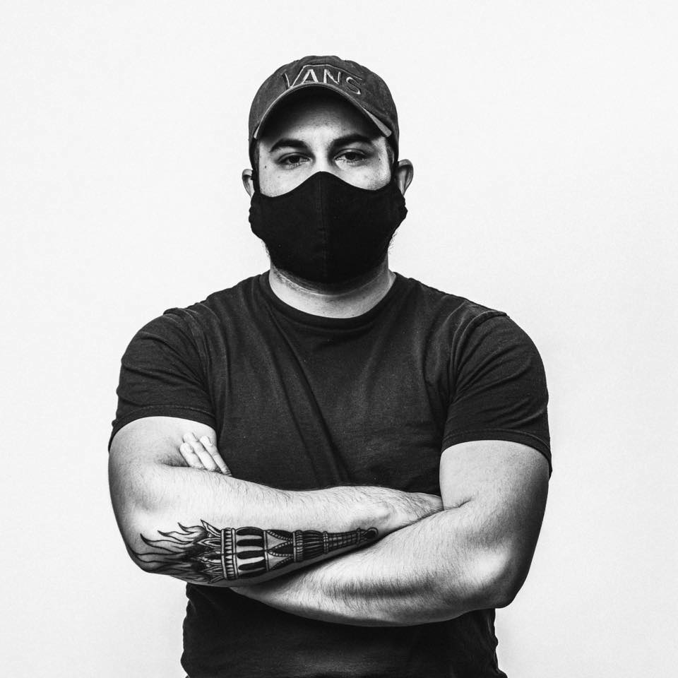 Una fotografía de Adrián Orozco a blanco y negro. Porta una gorra y un cubrebocas. Tiene sus brazos cruzados.