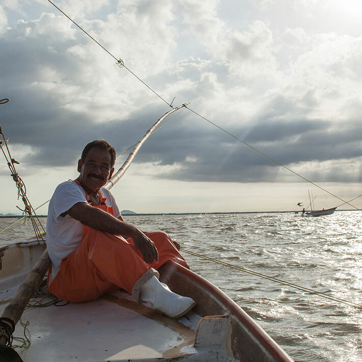 Un hombre de SUCEDE en una panga con el mar de fondo. Viste camiseta blanca y overall de trabajo naranja.