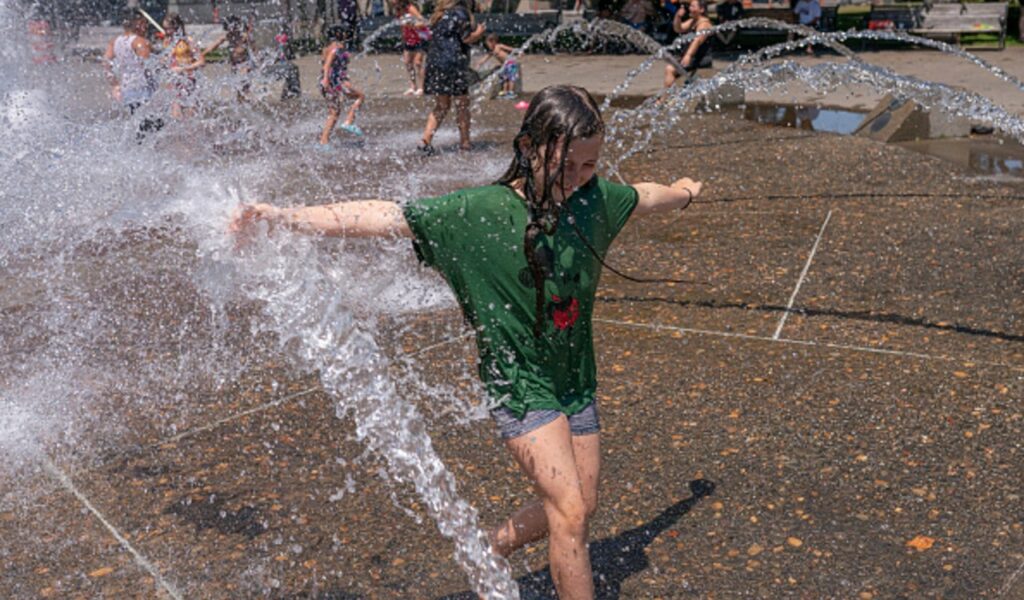 Una niña se moja con el agua de una fuente que emano chorros desde el suelo.